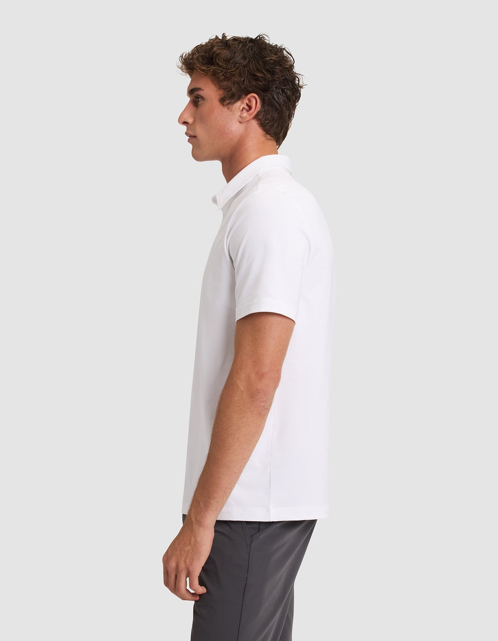 A young man stands in profile against a plain light gray background, wearing the Cozy Earth Men's Everyday Polo in white and gray pants. |Color:White