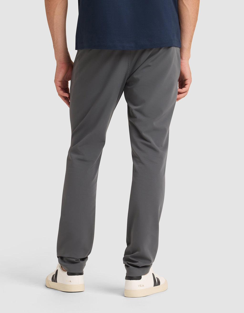 A person stands facing away, wearing Cozy Earth's Men's Everywhere Pant in dark gray, a navy blue shirt, and white sneakers with black heel accents against a plain light background.  |Color:Coal