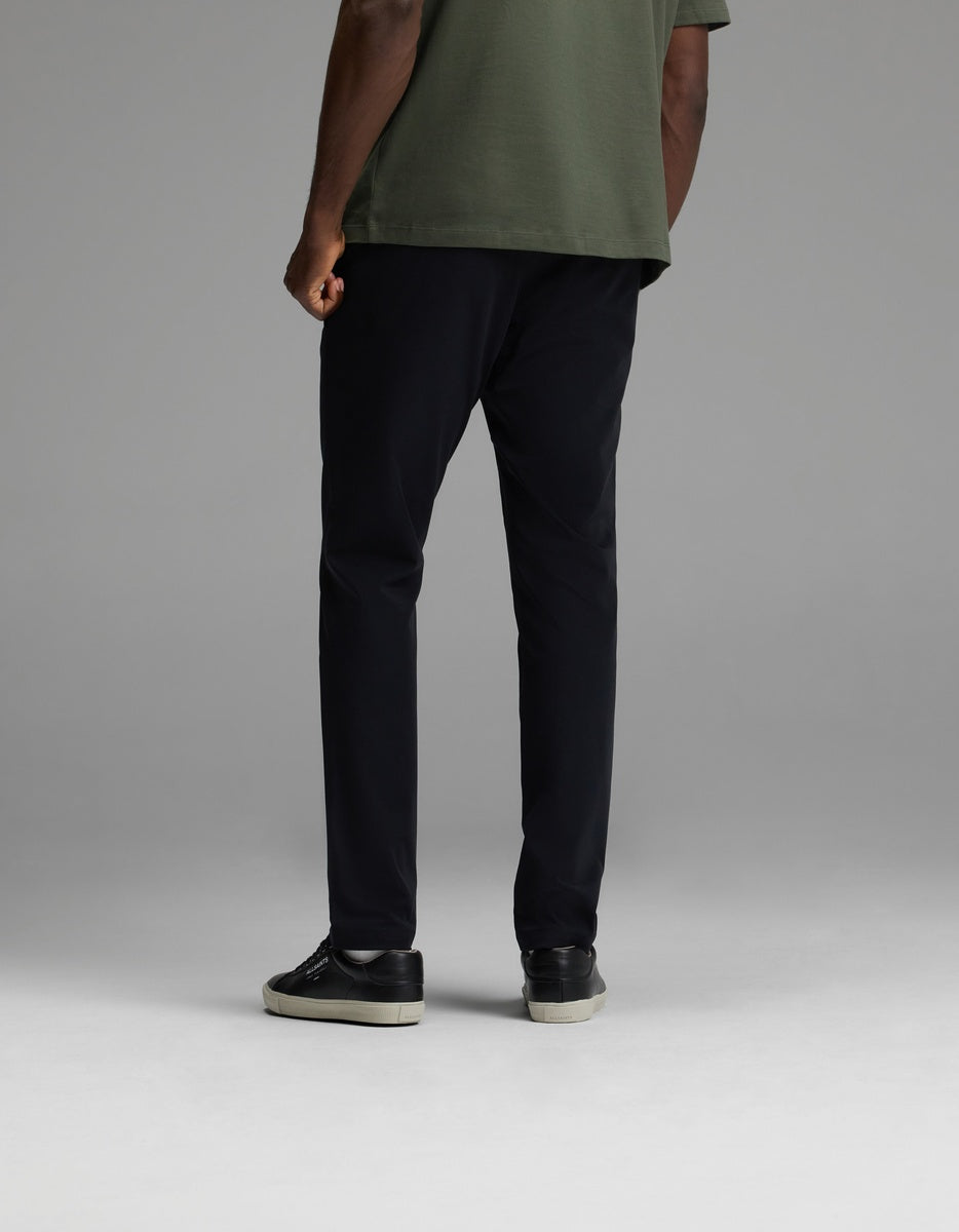 A person stands facing away against a plain gray background, wearing black Cozy Earth Men’s Everywhere Pants, paired with a green short-sleeve shirt and black sneakers with white soles.|Color:Jet Black