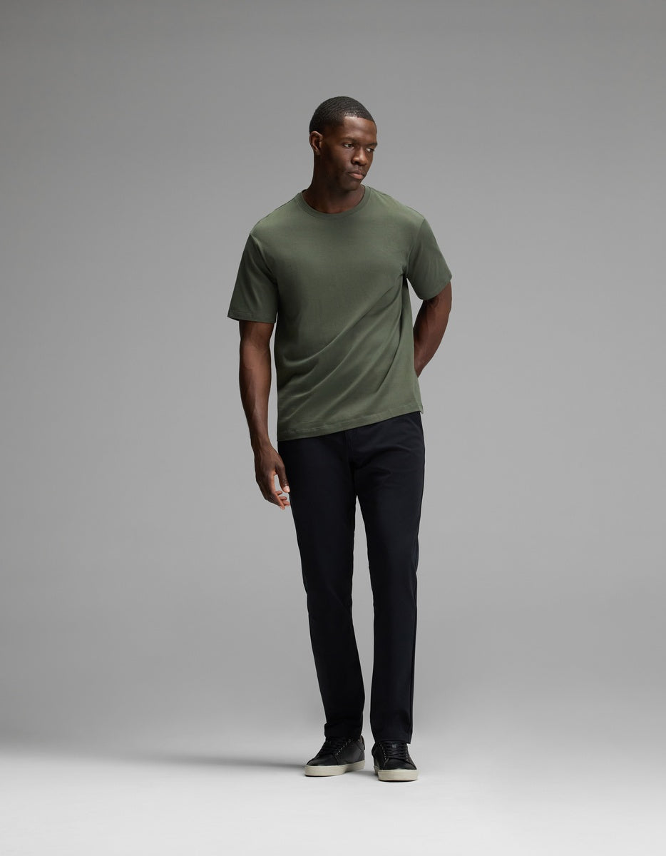 A man stands in a studio wearing Cozy Earth’s Men’s Everywhere Pant, paired with a green t-shirt and black shoes. He faces slightly to his left with one hand in his pocket against a plain gray background.|Color:Jet Black