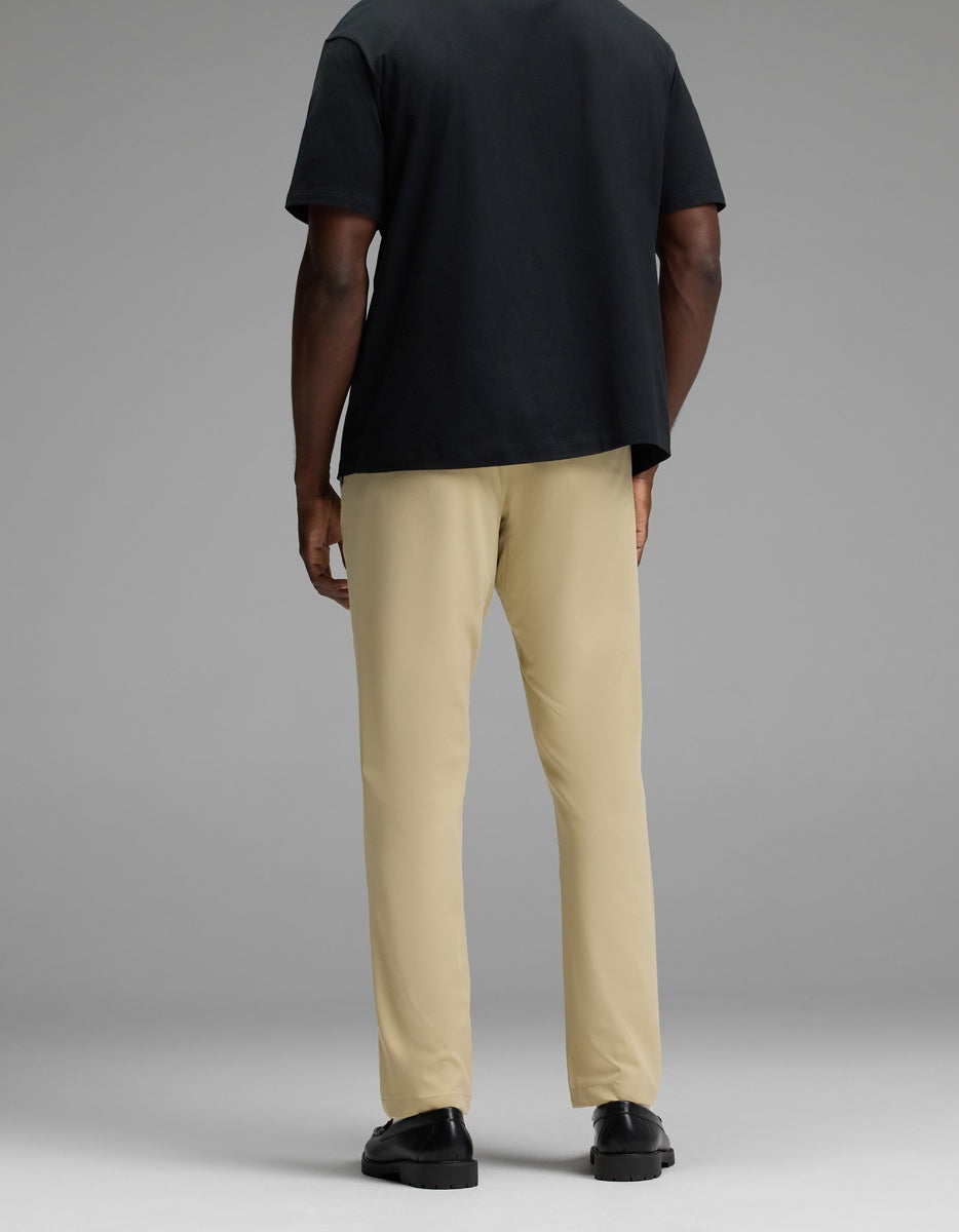 A person stands facing away, wearing Cozy Earth’s Men’s Everywhere Pant in beige, a black t-shirt, and black shoes against a plain gray background.|Color:Khaki