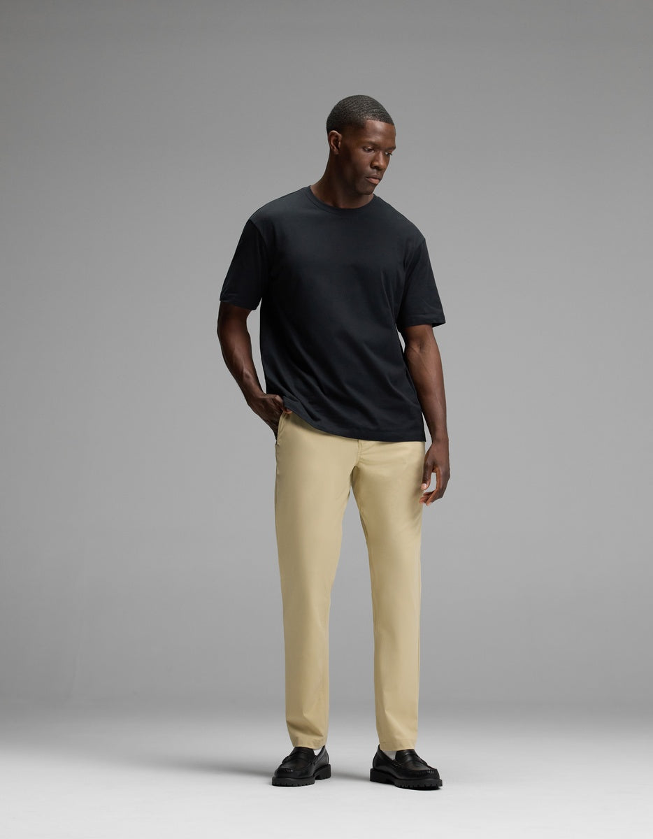 A man stands against a gray background, wearing Cozy Earth’s Men’s Everywhere Pant in beige, paired with a plain black t-shirt and black shoes. He looks down with one hand in his pocket.|Color:Khaki