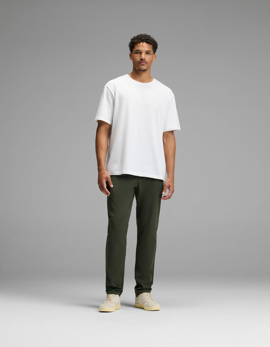 A man stands facing forward against a simple gray background, wearing olive green Men’s Everywhere Pant by Cozy Earth, a plain white t-shirt, and beige sneakers.|Color:Olive