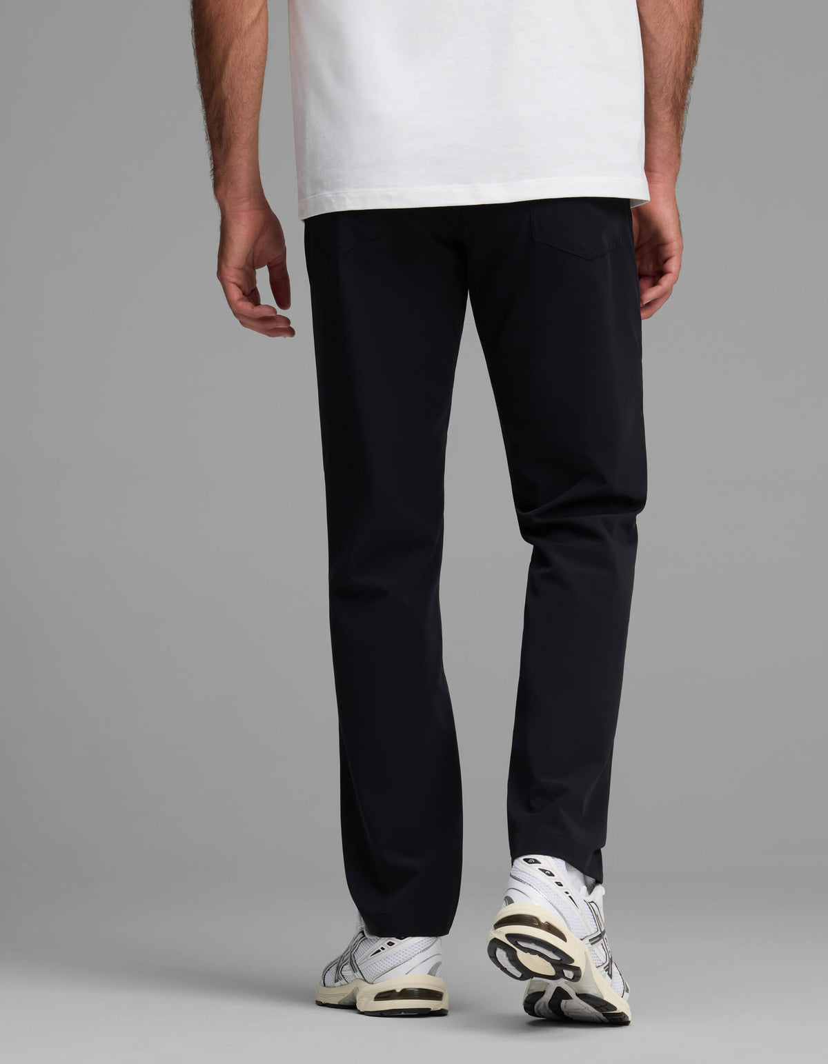 A person stands with their back to the camera against a plain gray background, wearing Cozy Earth’s Men's Everywhere Pant - Relaxed Fit, paired with a white T-shirt and white sneakers.|Color:Jet Black