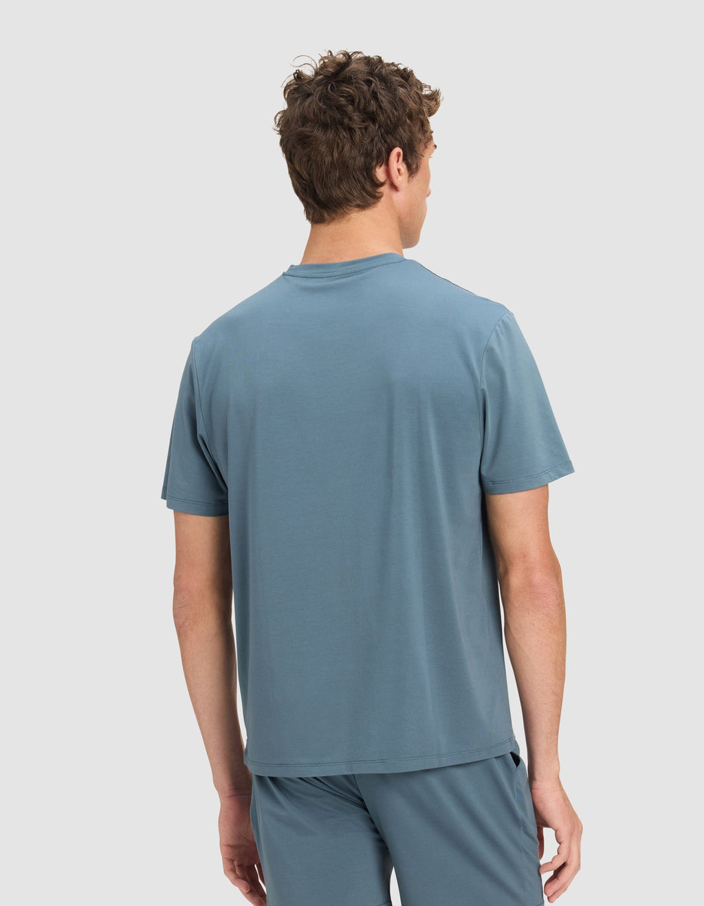 A man with short brown hair, seen from behind, is wearing the Cozy Earth Men's First Layer Pajama Short Sleeve Tee in light blue, paired with matching shorts against a plain light background.|Color:Dawn