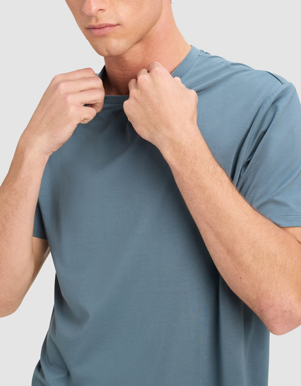 A person in a light blue Cozy Earth Men's First Layer Pajama Short Sleeve Tee holds the neckline with both hands. Their face is partially visible and neutral, set against a plain light background.|Color:Dawn