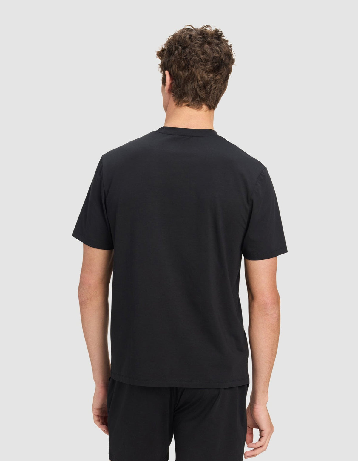 A man with curly brown hair stands against a light gray background, facing away from the camera, wearing a black Men's First Layer Pajama Short Sleeve Tee by Cozy Earth and black shorts.|Color:Jet Black