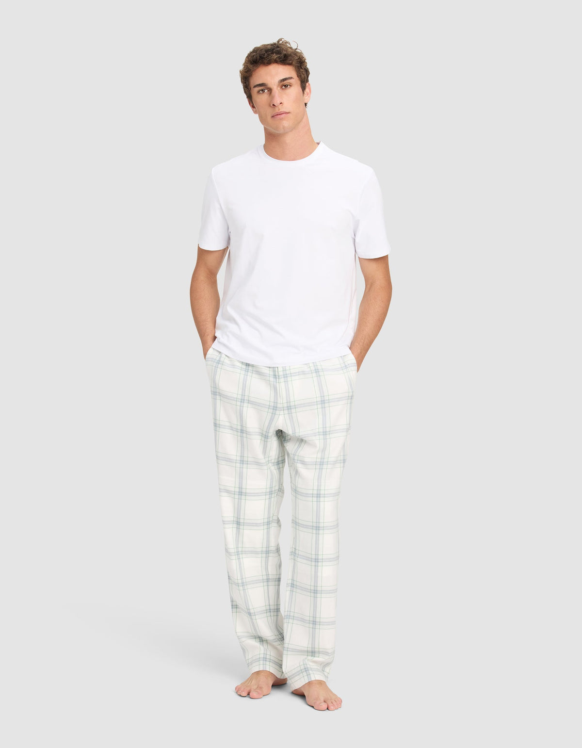 A young man stands barefoot against a plain background, wearing the Cozy Earth Men's First Layer Pajama Short Sleeve Tee and white plaid pajama pants, with his hands in his pockets.|Color:White