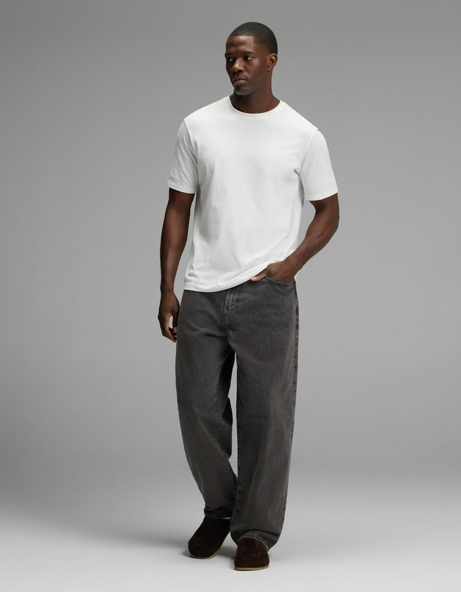 A man stands with one hand in his pocket against a plain gray background, wearing loose-fitting dark gray jeans, a plain white t-shirt, and Cozy Earth’s Lakehouse Clog in Suede.|Color:Espresso