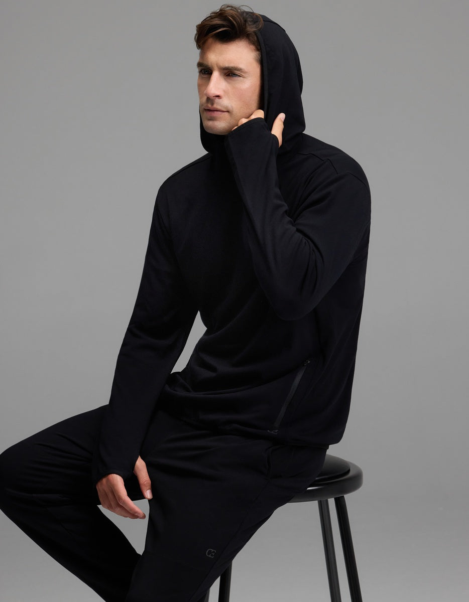 A man wearing the Cozy Earth Men’s Recovery Hoodie and matching pants sits on a black stool against a gray background, with one hand touching his hoodie near his chin.|Color:Black