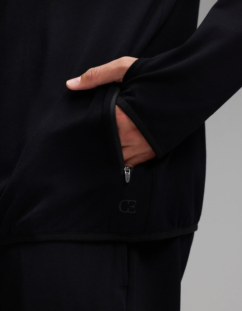 A person wearing the Cozy Earth Men’s Recovery Hoodie in black, with their hand partially inside a zippered pocket. The hoodie features subtle piping along the edges and a small Cozy Earth logo near the bottom of the pocket.|Color:Black