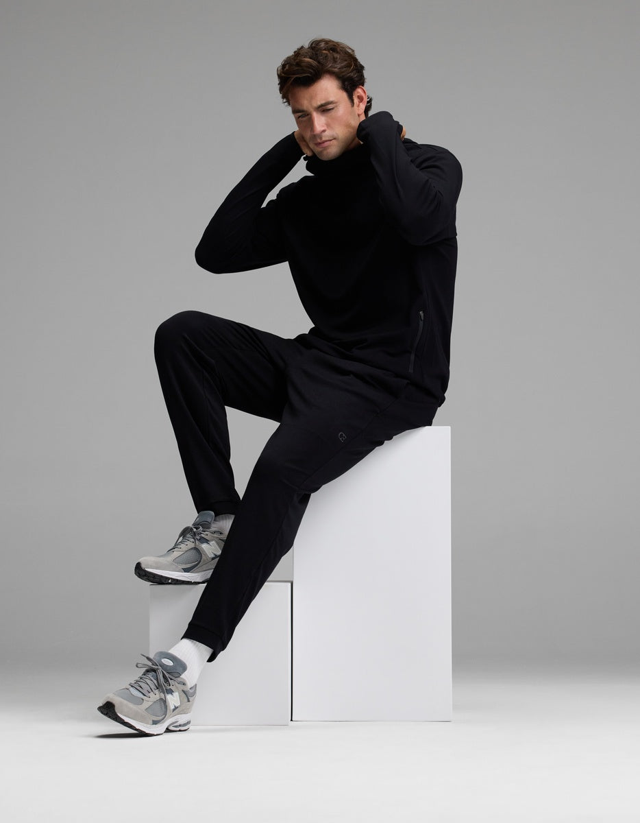 A man wearing Cozy Earth’s Men’s Recovery Jogger and gray sneakers sits on white blocks with one knee raised, hands behind his neck, looking downward against a plain gray background.|Color:Black