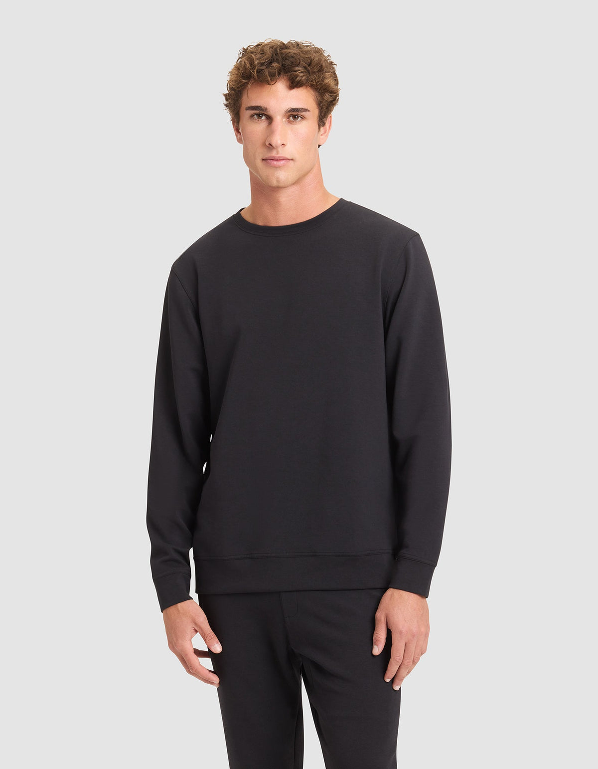 A young man with curly brown hair wears the Cozy Earth Men's StretchTech Crewneck in black with matching black pants, standing against a light gray background. |Color:Jet Black