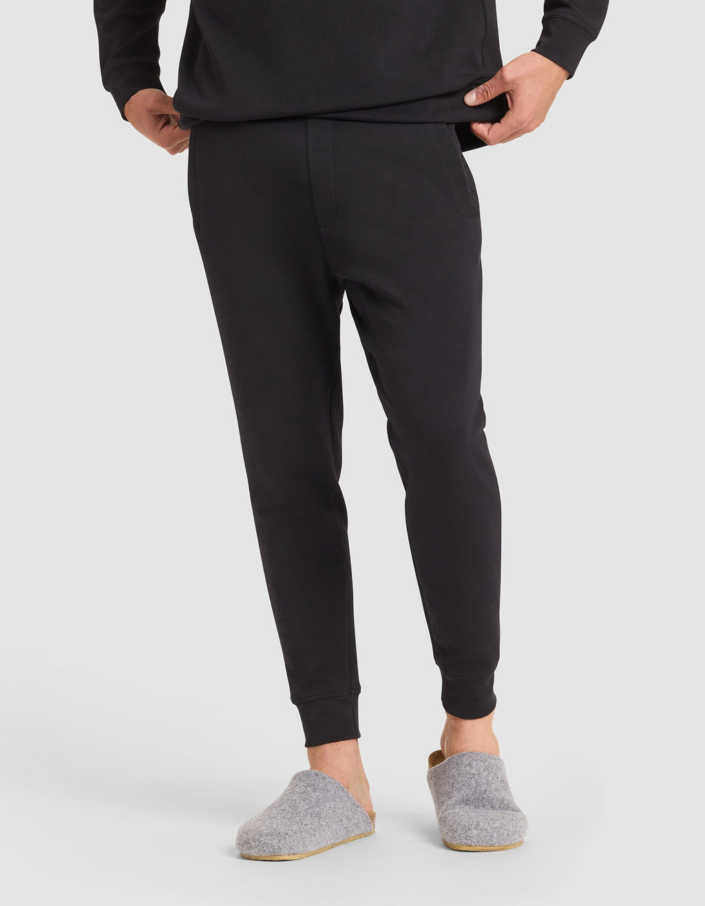 A person stands on a white background, hands in pockets, wearing Cozy Earth Men's StretchTech Jogger and a black long-sleeve top, paired with gray slip-on house slippers.  |Color:Jet Black