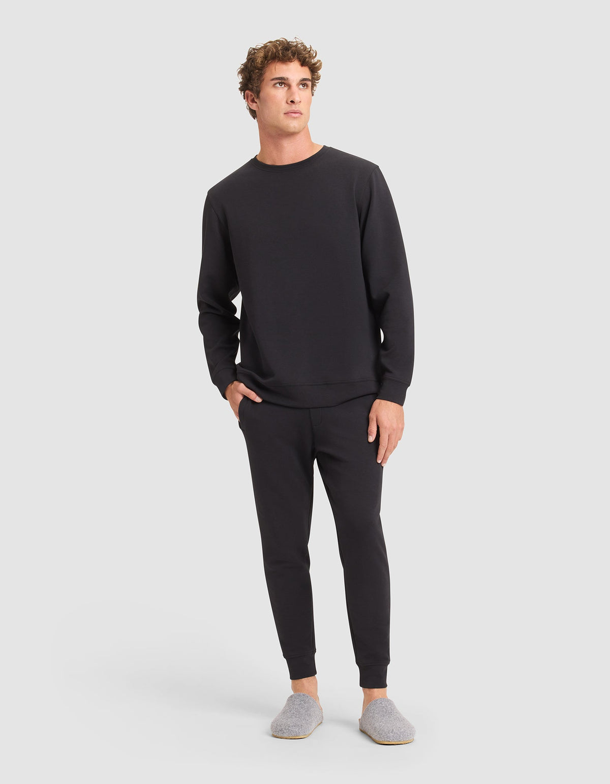 A man stands against a plain background wearing Cozy Earth’s Men's StretchTech Jogger in black, paired with a black sweatshirt and gray slippers. He has one hand in his pocket and looks slightly to the side. |Color:Jet Black