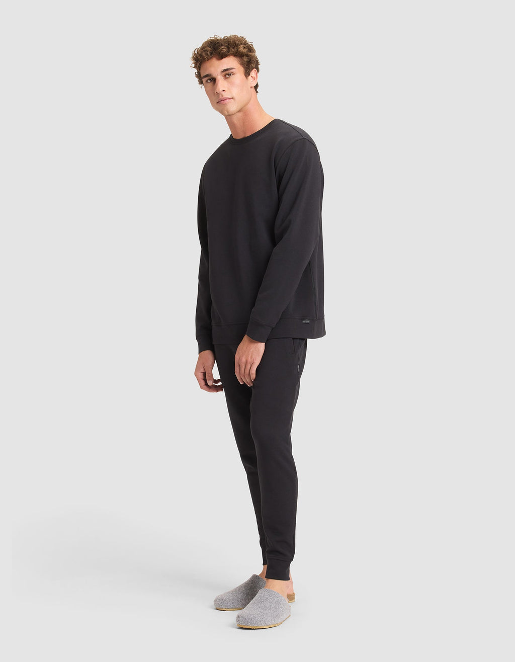 A young man models the Cozy Earth Men's StretchTech Crewneck & Jogger Set in black, paired with light gray slippers, standing against a plain light gray background and gazing slightly sideways at the camera. |Color:Jet Black
