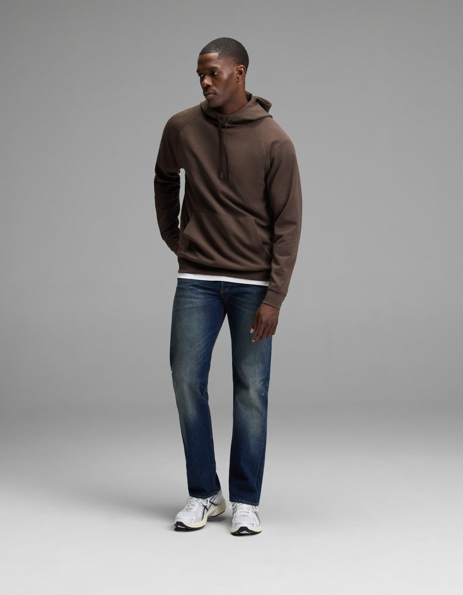 A person stands in a studio wearing the Cozy Earth Men’s StretchTech Hoodie, paired with blue jeans and white sneakers. They look down and to the side with their hands relaxed by their sides against a plain gray background.|Color:Cacao