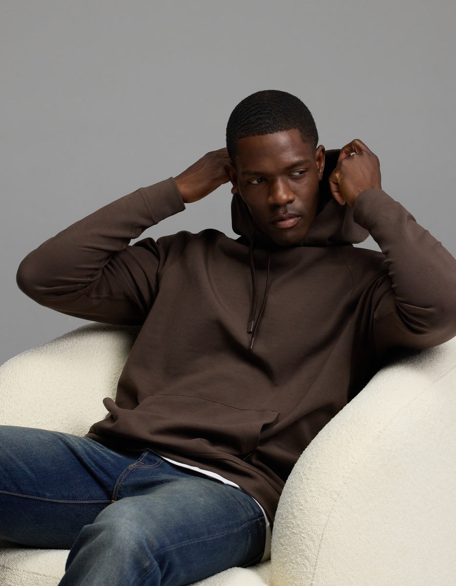 A man wearing the Cozy Earth Men’s StretchTech Hoodie in brown and blue jeans sits on a white chair, adjusting his hood with both hands while looking slightly to the side against a plain gray background.|Color:Cacao
