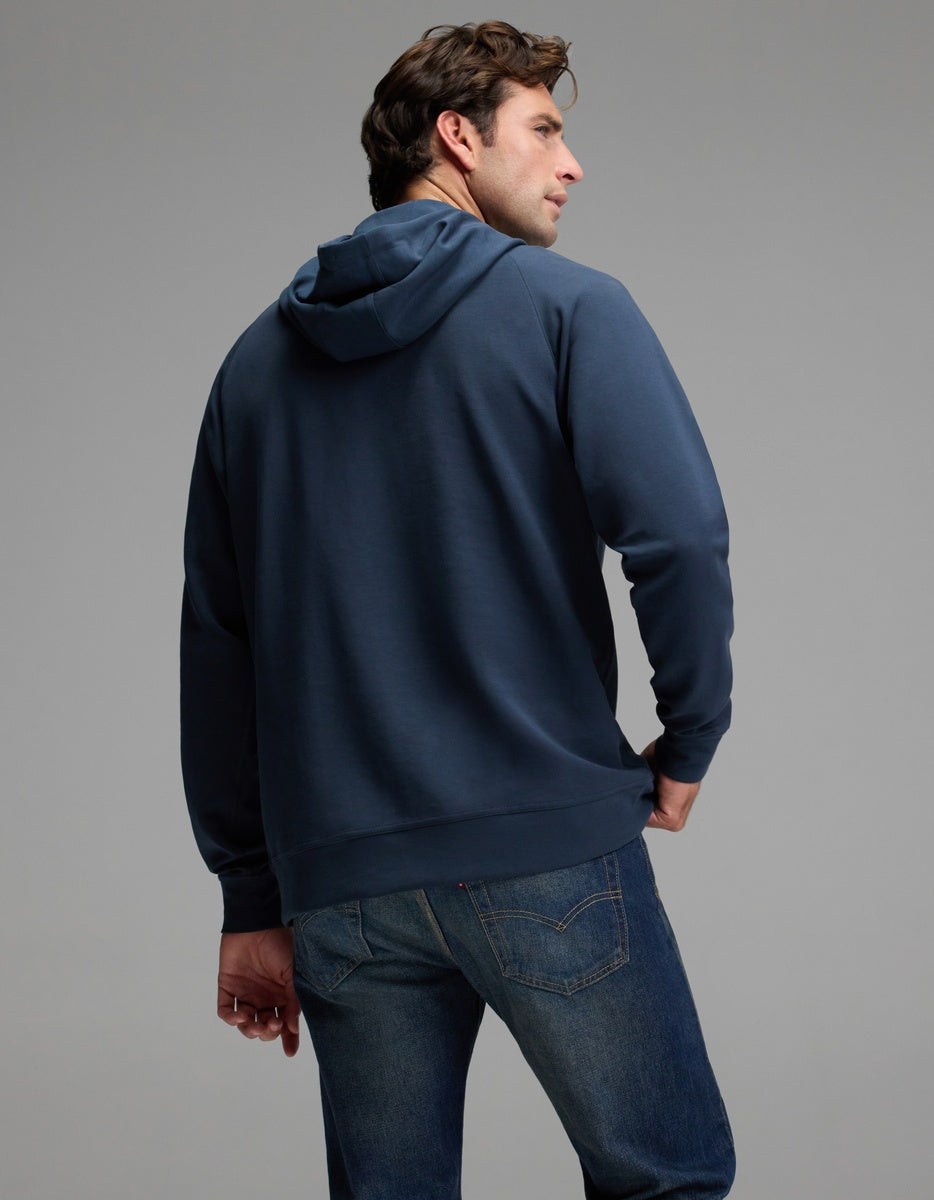 A man wearing the Cozy Earth Men’s StretchTech Hoodie in blue and a pair of jeans stands with his back to the camera, head turned slightly to the side, set against a plain gray background.|Color:Eclipse