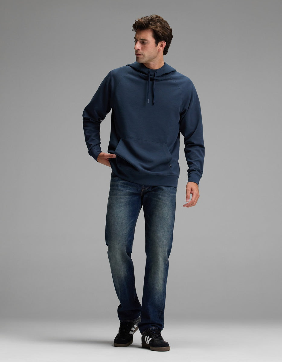 A man wearing the Cozy Earth Men’s StretchTech Hoodie, blue jeans, and black sneakers walks against a light gray background, looking to his right with one hand in his pocket.|Color:Eclipse