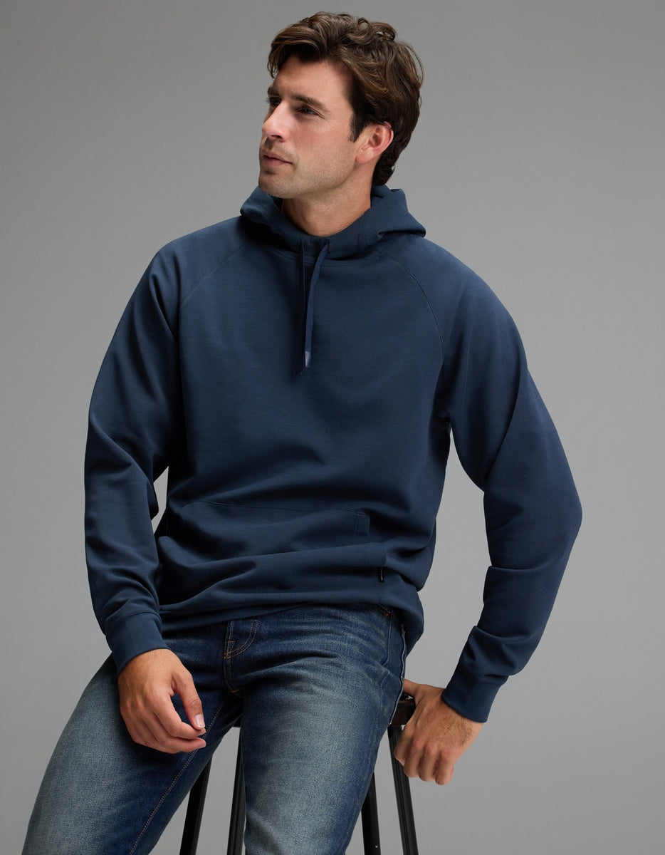 A man wearing the Cozy Earth Men’s StretchTech Hoodie in dark blue and jeans sits on a stool against a plain gray background, looking slightly to the side.|Color:Eclipse