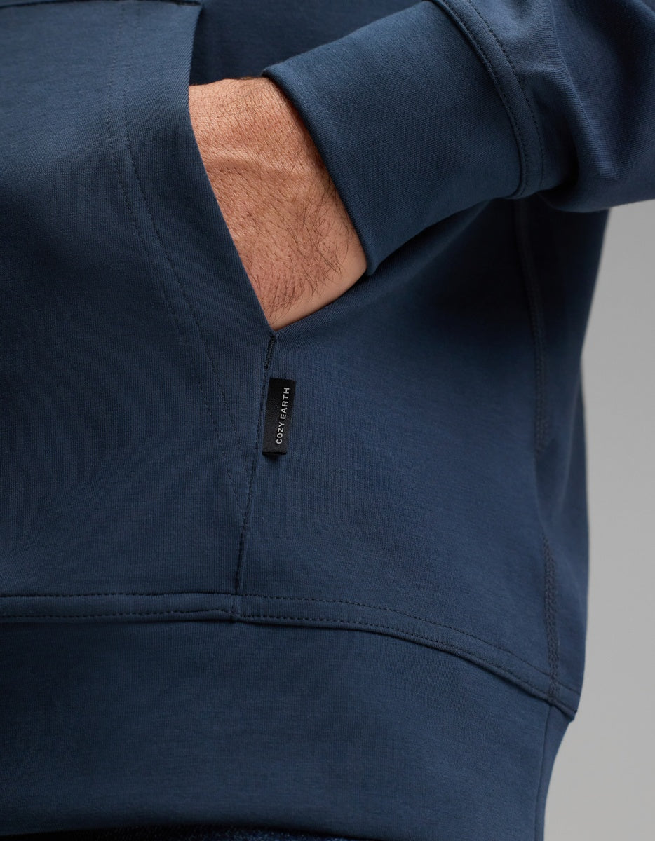 A close-up of a person’s hand partially tucked into the pocket of a dark blue Cozy Earth Men’s StretchTech Hoodie, featuring a small black fabric tag on the seam.|Color:Eclipse