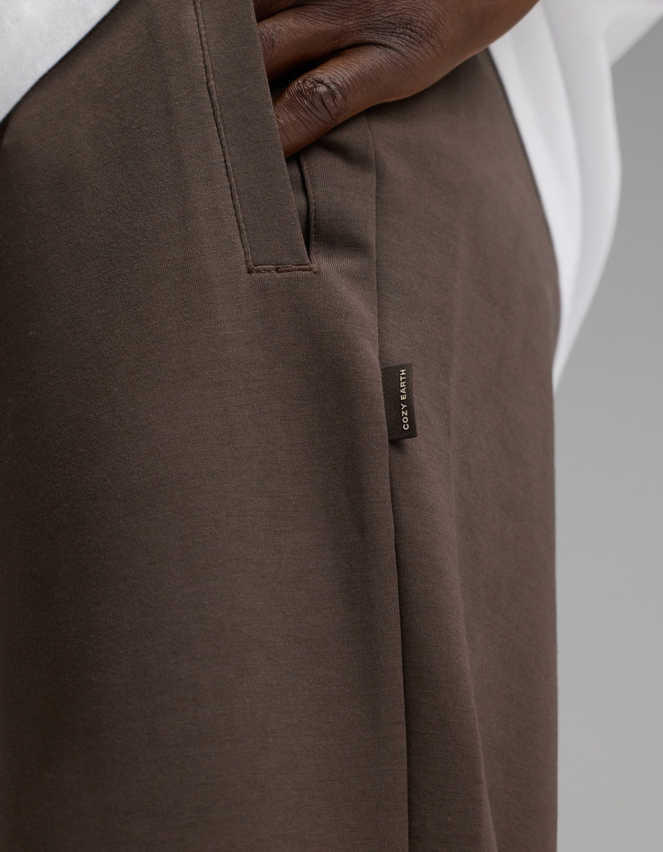 A close-up of a person’s hand in the pocket of Cozy Earth Men’s StretchTech Jogger pants in brown, featuring a small fabric tag with text. The person is also wearing a white shirt.|Color:Cacao