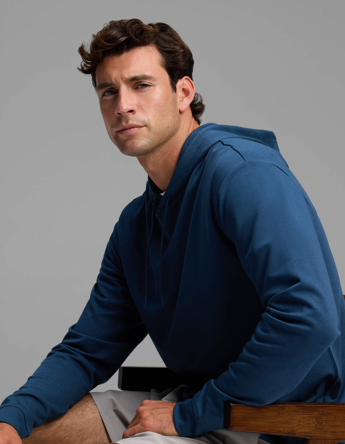 A man with short brown hair wears the Men's Versa Hoodie by Cozy Earth in blue and light gray shorts as he sits on a wooden chair, gazing slightly to the side with a neutral expression against a plain gray background.|Color:Obsidian