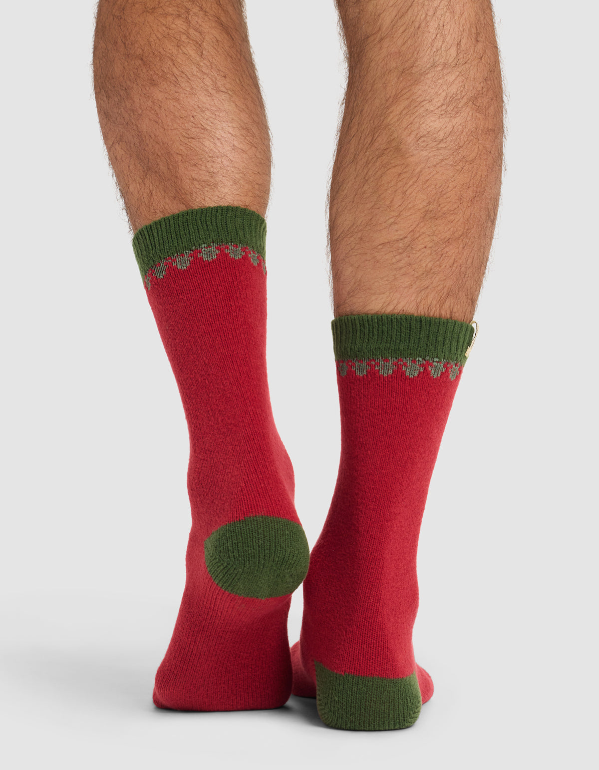 Close-up of a person's legs wearing The Mistletoe Plush Lounge Sock by Cozy Earth—red socks with green heels, toes, and cuffs—standing on a plain light background.|Color:Cranberry