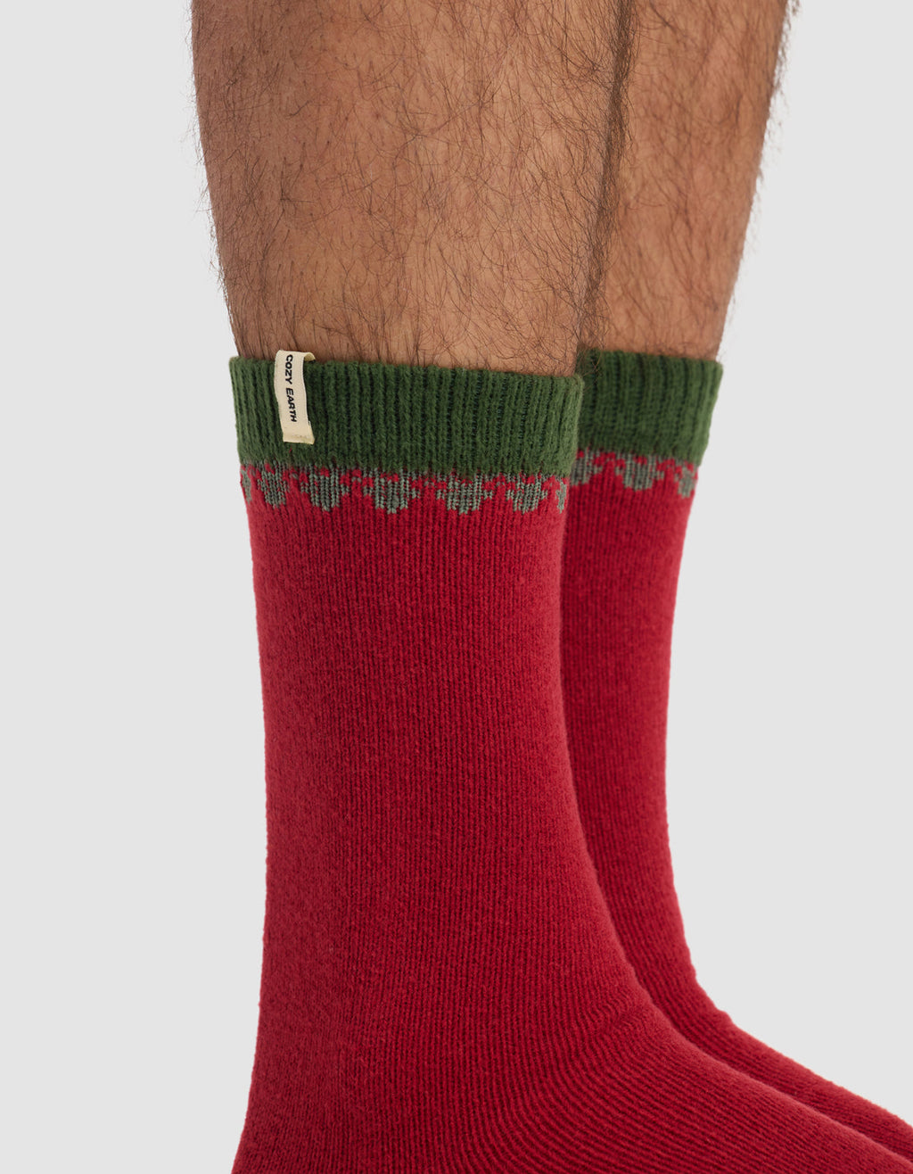 Close-up of a person's lower legs wearing The Mistletoe Plush Lounge Sock by Cozy Earth—red socks with green ribbed cuffs and a small white fabric tag—against a plain light background.|Color:Cranberry