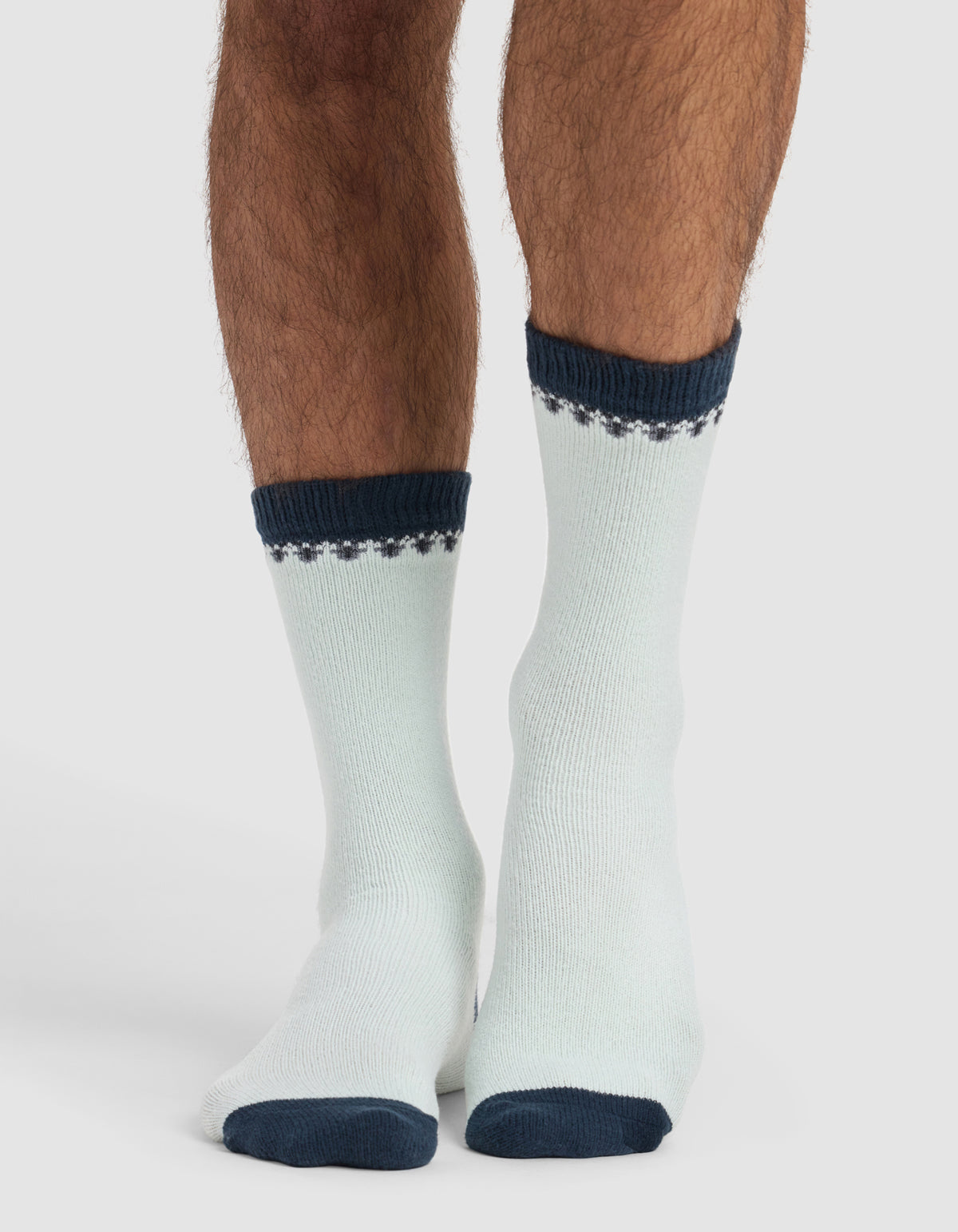 A person wears The Mistletoe Plush Lounge Sock by Cozy Earth: light-colored with dark blue toes, heels, cuffs, and a patterned band near the top. The background is plain and light.|Color:Mist