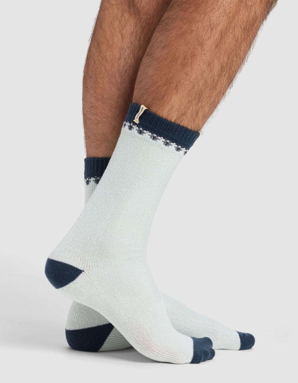 Someone stands on a white background wearing The Mistletoe Plush Lounge Sock by Cozy Earth, featuring light colors with dark blue toes, heels, and cuffs, plus a subtle decorative pattern near the top.|Color:Mist