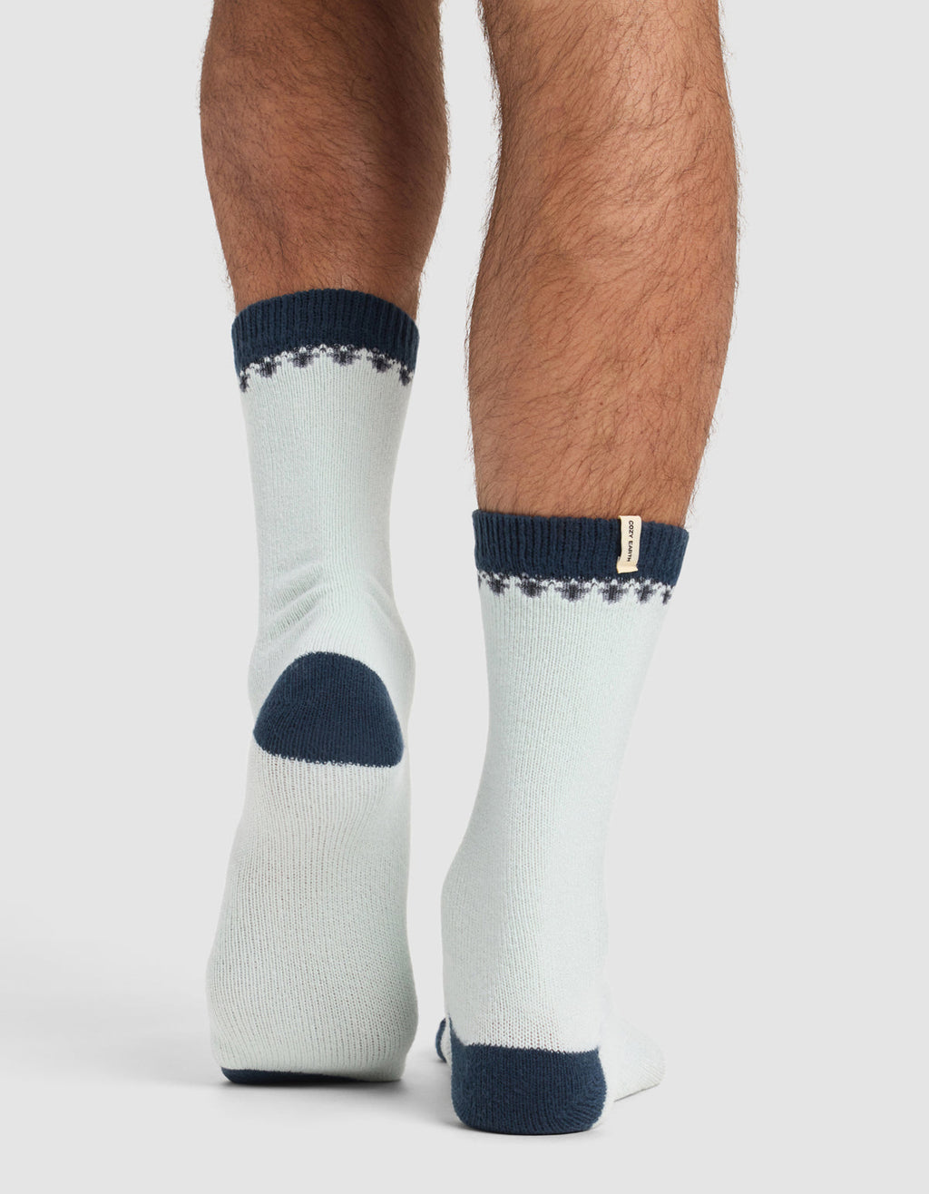 A person stands on tiptoe, viewed from behind, wearing Cozy Earth’s The Mistletoe Plush Lounge Sock—light-colored with dark blue heels, toes, cuffs, and a dotted pattern near the top.|Color:Mist