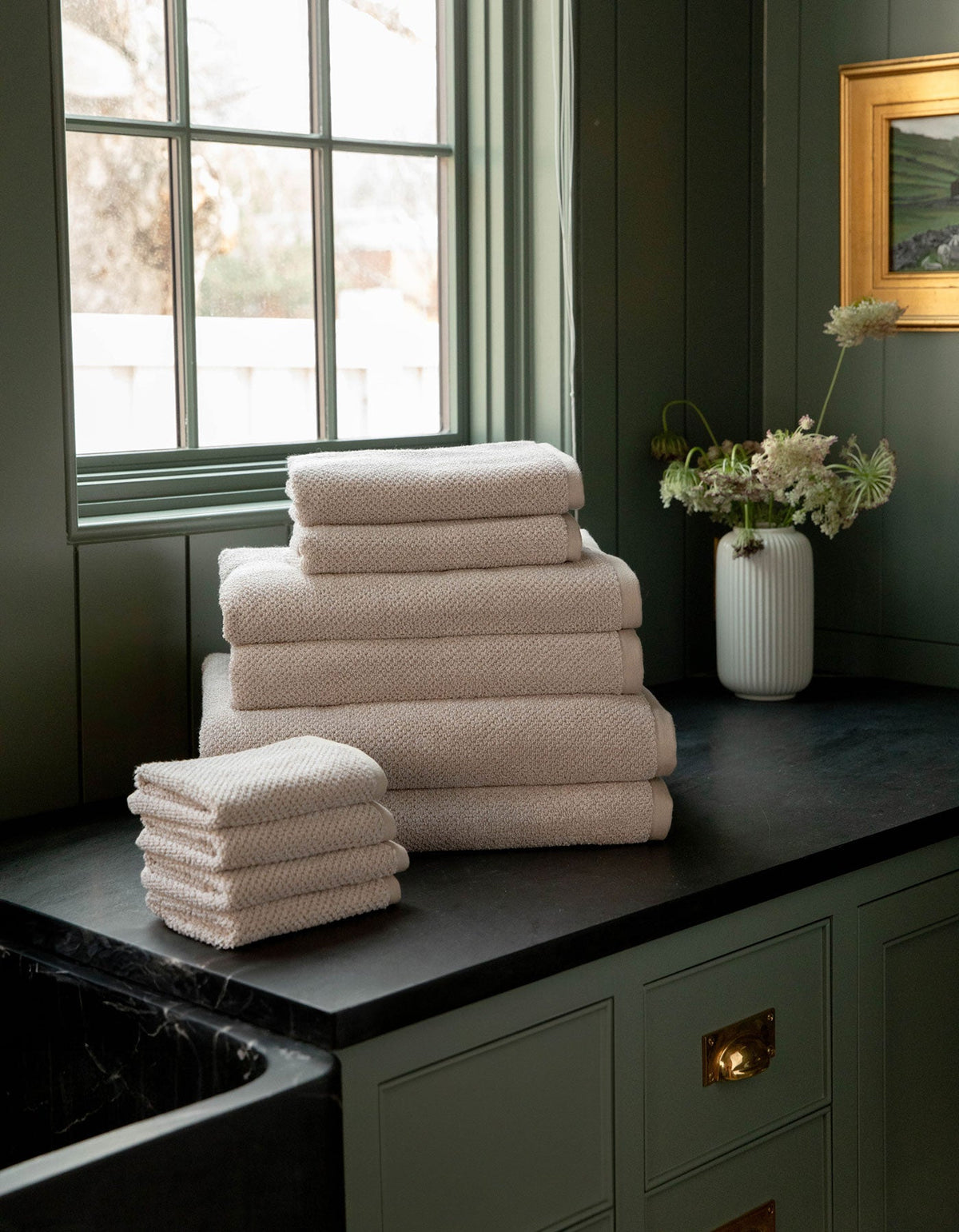 The Complete Nantucket Bath Bundle by Cozy Earth, in beige, is neatly stacked on a black countertop near a window with a vase of flowers and pale green cabinetry in the background.|Color:Heathered Sand