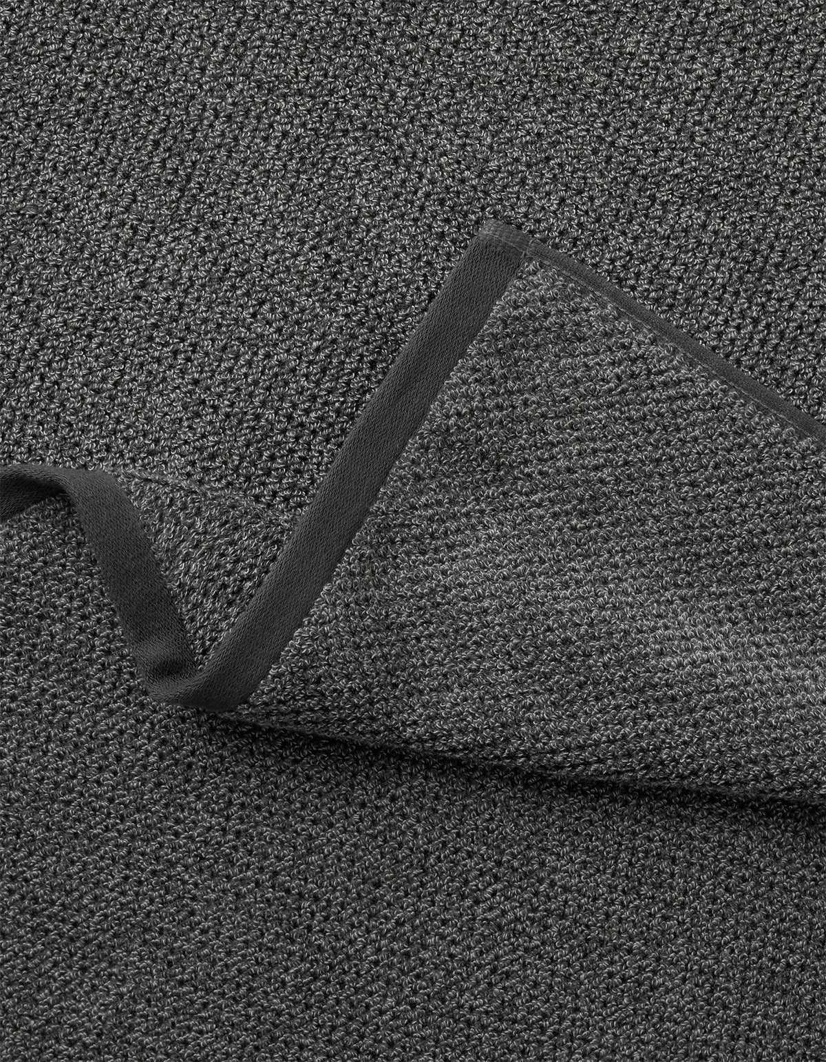 A close-up of Cozy Earth's Nantucket Bath Towel Set shows its textured dark gray fabric with a folded corner, revealing the same woven material on both sides and a smooth, dark border along the edge.|Color:Graphite