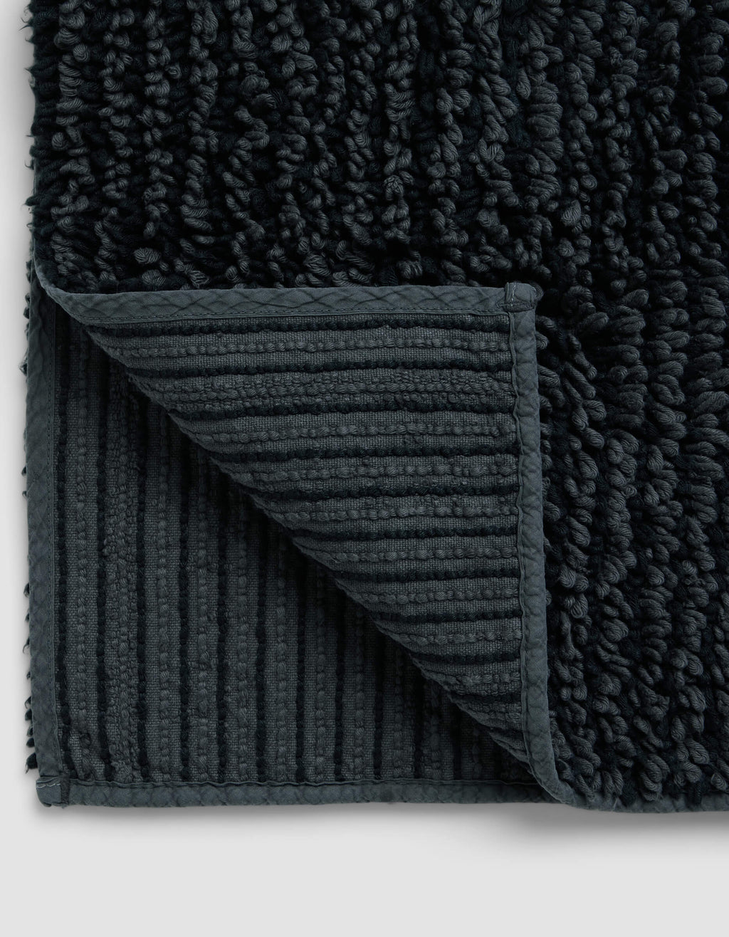 Close-up of the Cozy Earth Plush Loop Bath Mat with a corner folded, highlighting its ribbed, non-slip underside and thick, soft plush pile on top.|Color:Graphite