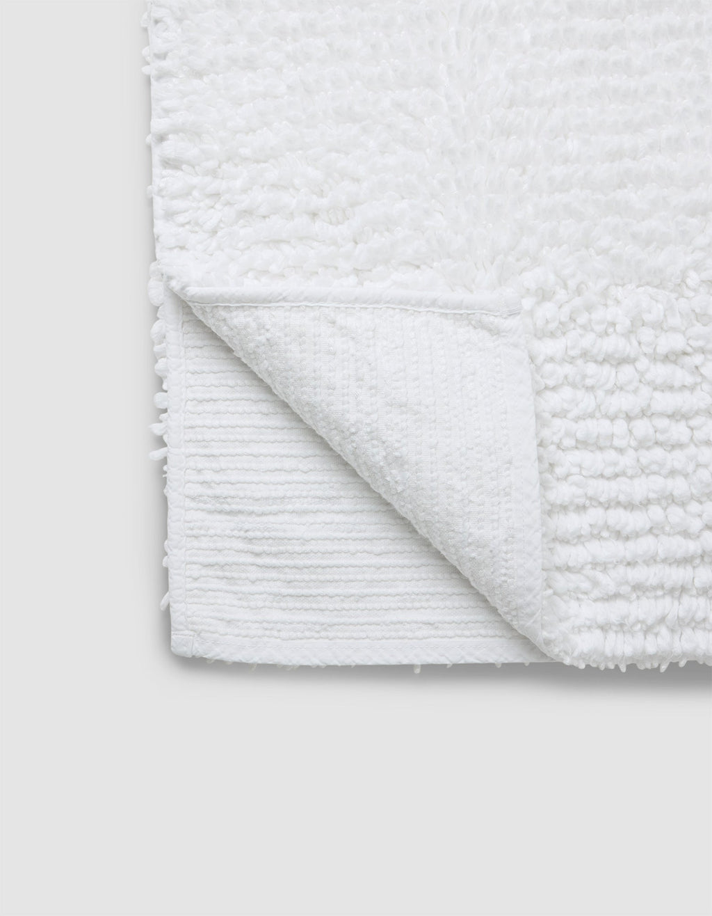 A close-up of the Cozy Earth Plush Loop Bath Mat in white, featuring a soft, textured surface with one corner flipped to show its flat underside. The mat is set against a plain light gray background.|Color:White