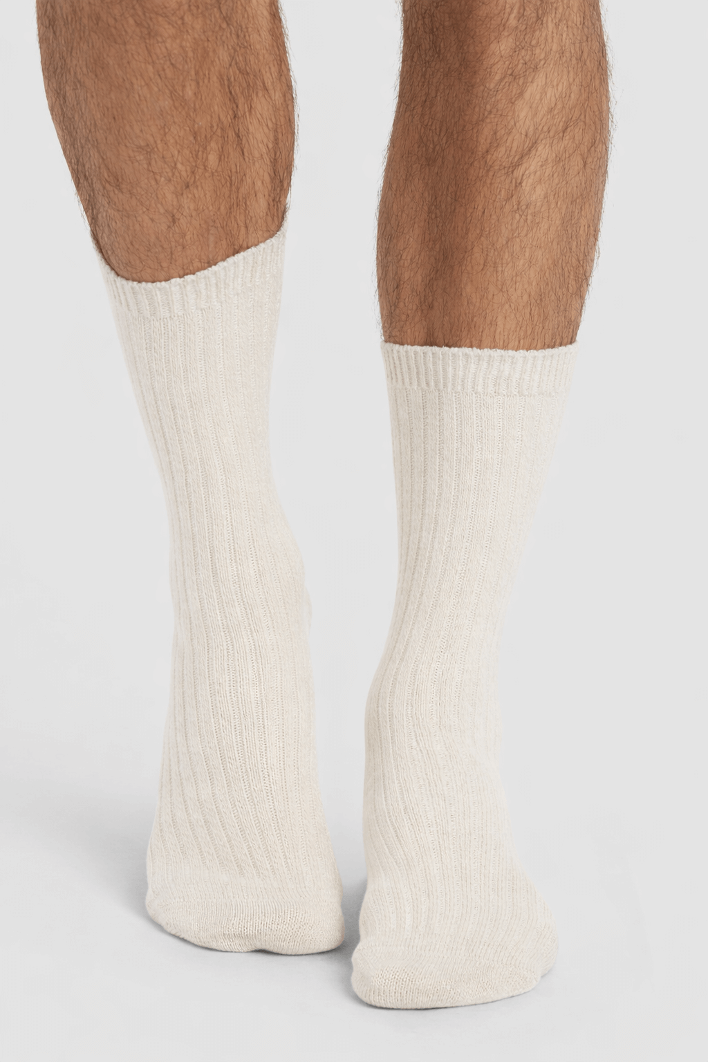 A close-up of a person's lower legs wearing Cozy Earth's Men's Plush Lounge Calf Sock 3-Pack in cream, ribbed design, standing on a white surface.|Color:Cacao/Taupe/Alabaster