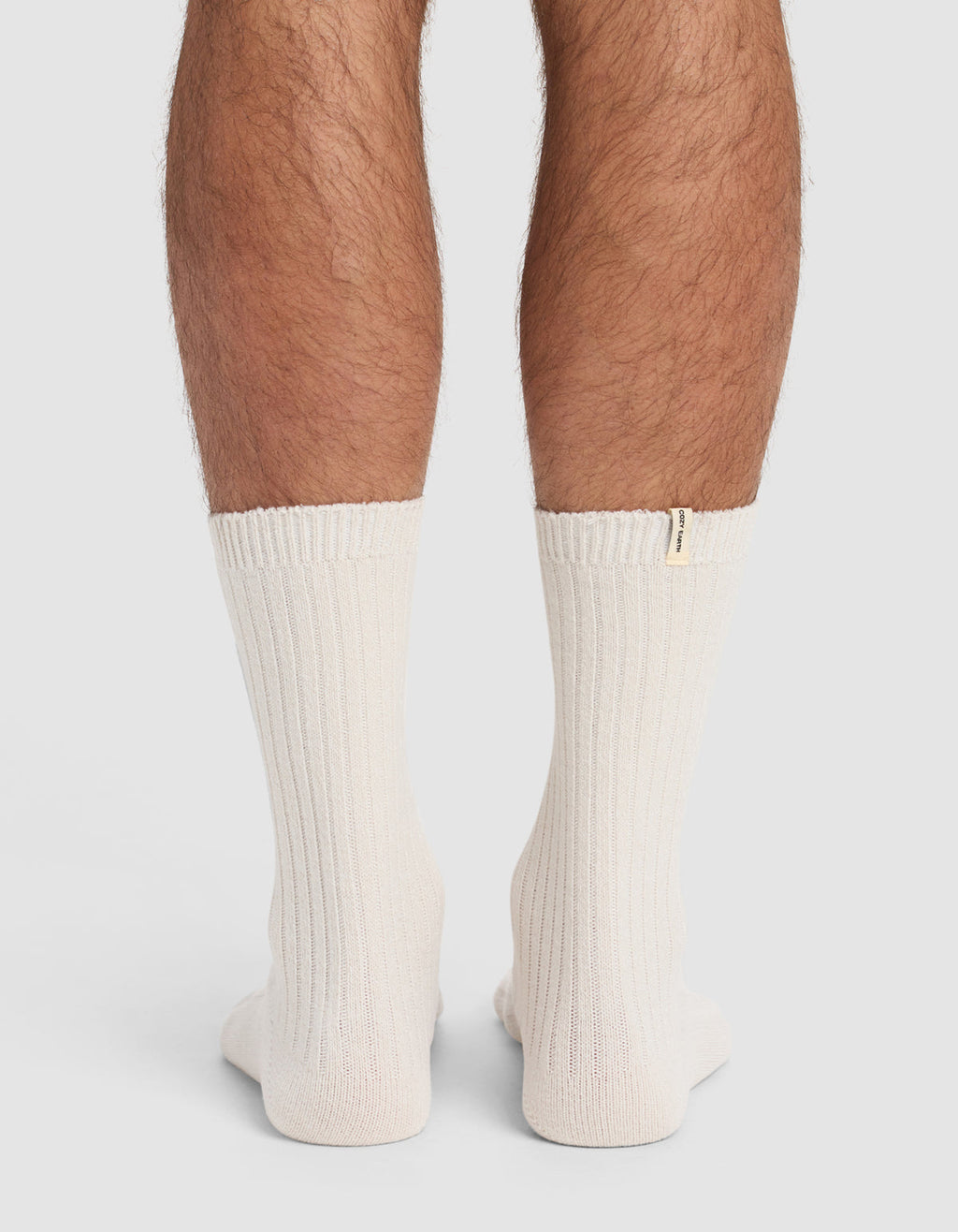 Back view of a person's lower legs wearing The Plush Lounge Calf Sock 3-Pack by Cozy Earth—thick, ribbed, off-white socks with a small beige tag near the top edge. Plain, light-colored background.|Color:Cacao/Taupe/Alabaster