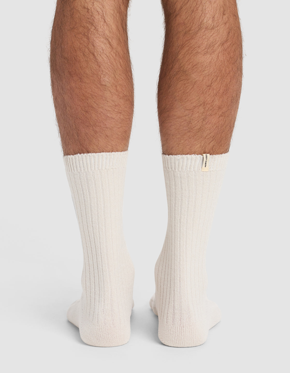Back view of a person's lower legs wearing The Plush Lounge Calf Sock 3-Pack by Cozy Earth—thick, ribbed, off-white socks with a small beige tag near the top edge. Plain, light-colored background.|Color:Cacao/Taupe/Alabaster