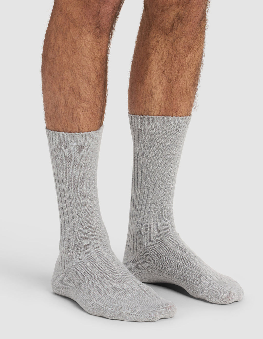 A person wears Cozy Earth's The Plush Lounge Calf Sock 3-Pack in light gray, ribbed crew style, with only their lower legs and feet visible against a plain white background.|Color:Charcoal/Coal/Stone
