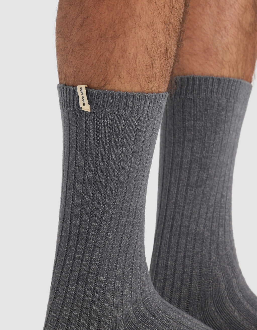 Close-up of a person's lower legs wearing The Plush Lounge Calf Sock in dark gray from Cozy Earth, featuring ribbed texture and a small beige tag near the top. Plain white background.|Color:Charcoal/Coal/Stone