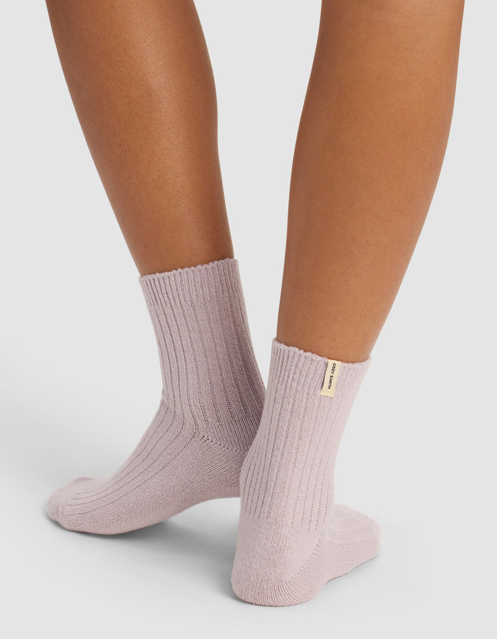 A person stands against a plain background, wearing The Plush Modern Crew Sock by Cozy Earth in light purple ribbed fabric with a small beige tag on one sock.|Color:Lavender