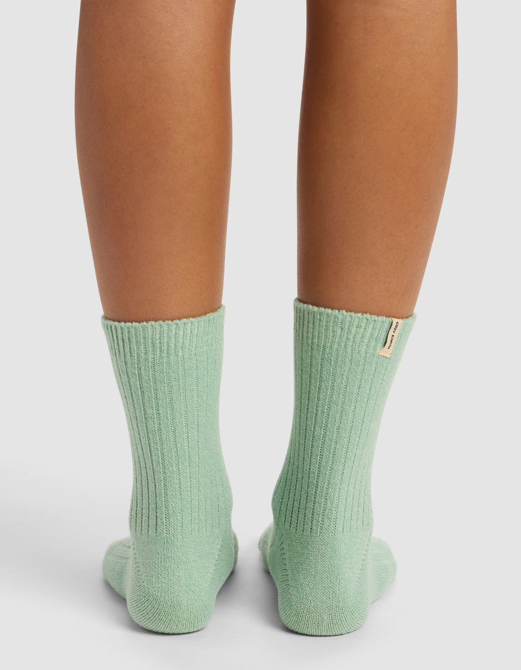 Only the lower legs and feet, seen from behind, show someone standing on a white surface wearing Cozy Earth’s The Plush Modern Crew Sock in mint green ribbed fabric that reaches mid-calf.|Color:Wintergreen