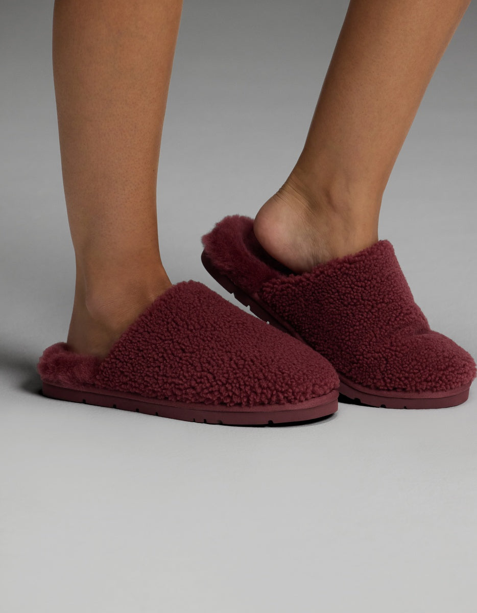 A person wearing Cozy Earth Puffy Sheep Slippers in dark red stands on a light gray surface. The slippers feature a plush, textured design with open heels.|Color:Mulberry