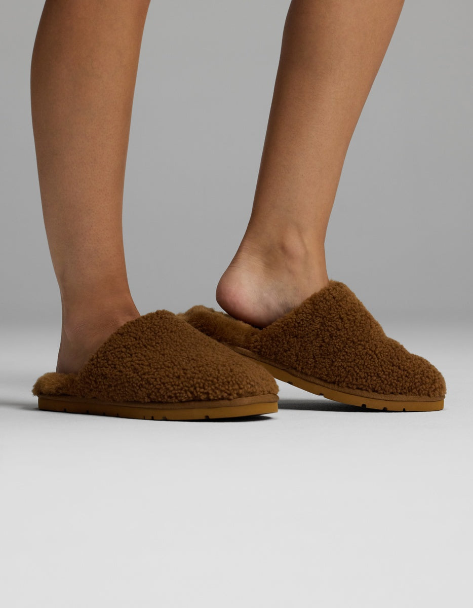 A person wearing Cozy Earth Puffy Sheep Slippers stands on a light grey surface, with one foot slightly raised to reveal the back of the slipper and the other resting flat on the ground.|Color:Teddy