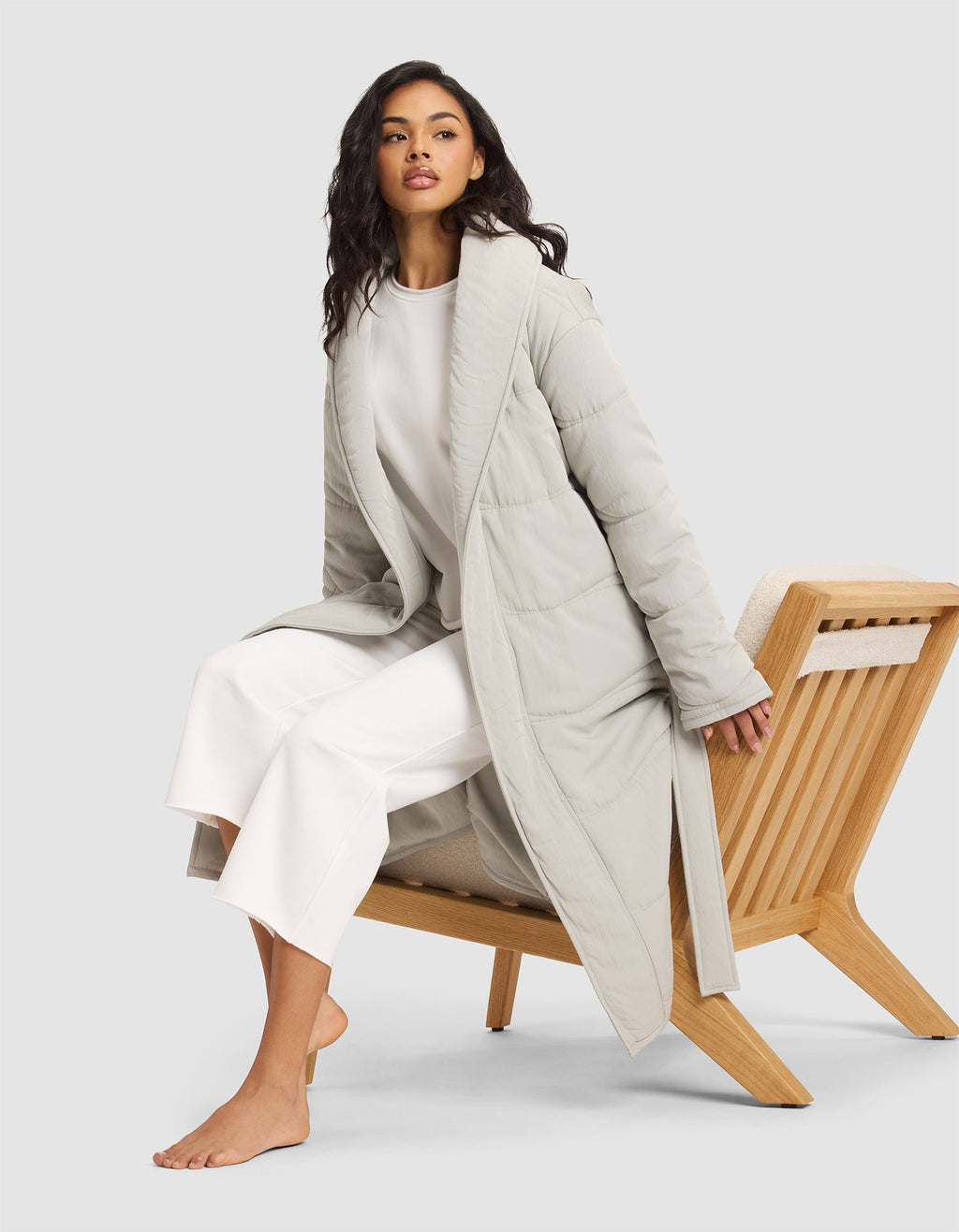 A woman with long dark hair sits barefoot on a wooden chair, wearing the Cozy Earth Women's Quilted House Coat over a white top and wide-legged white pants, against a plain light background. |Color:Aloe