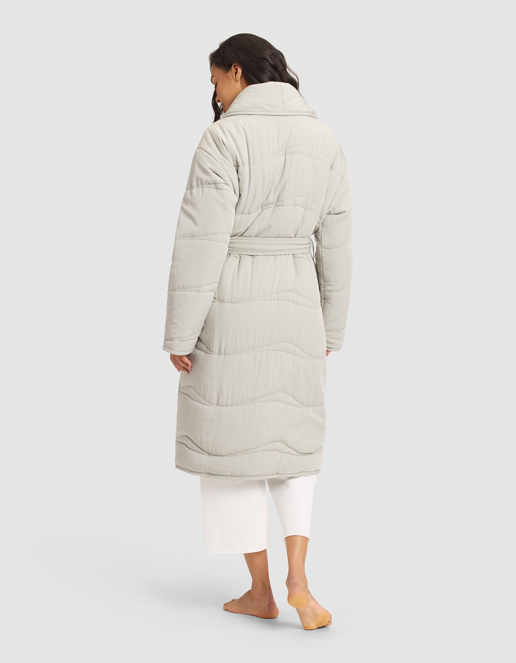 A woman with long, wavy dark hair models the Cozy Earth Women's Quilted House Coat in light gray, paired with white pants. She stands barefoot with her back to the camera against a plain background. |Color:Aloe