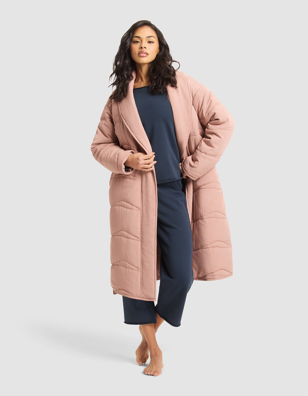 A woman with long wavy hair poses barefoot against a light gray background, wearing the Cozy Earth Women's Quilted House Coat in blush pink over navy blue loungewear. |Color:Rosewood