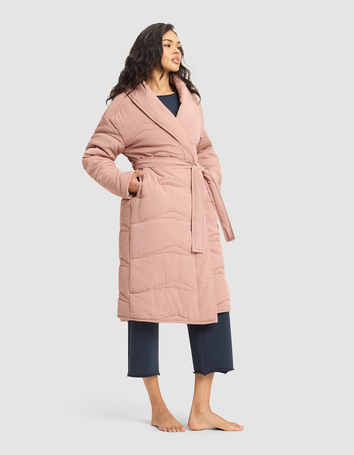A woman stands barefoot in the Cozy Earth Women's Quilted House Coat, a long, light pink belted robe, layered over navy blue pajama pants and top, set against a plain light background. |Color:Rosewood